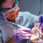 Dentist in protective gear performing a dental procedure using a drill on a patient&rsquo;s open mouth while wearing gloves and safety glasses.