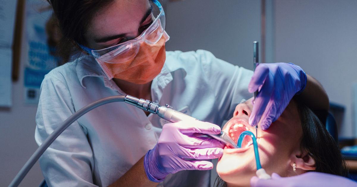MDW Centre - Complete Oral Care at a Dental Clinic in Aligarh - MDW Centre - MDW Centre Dentist in protective gear performing a dental procedure using a drill on a patient’s open mouth while wearing gloves and safety glasses.