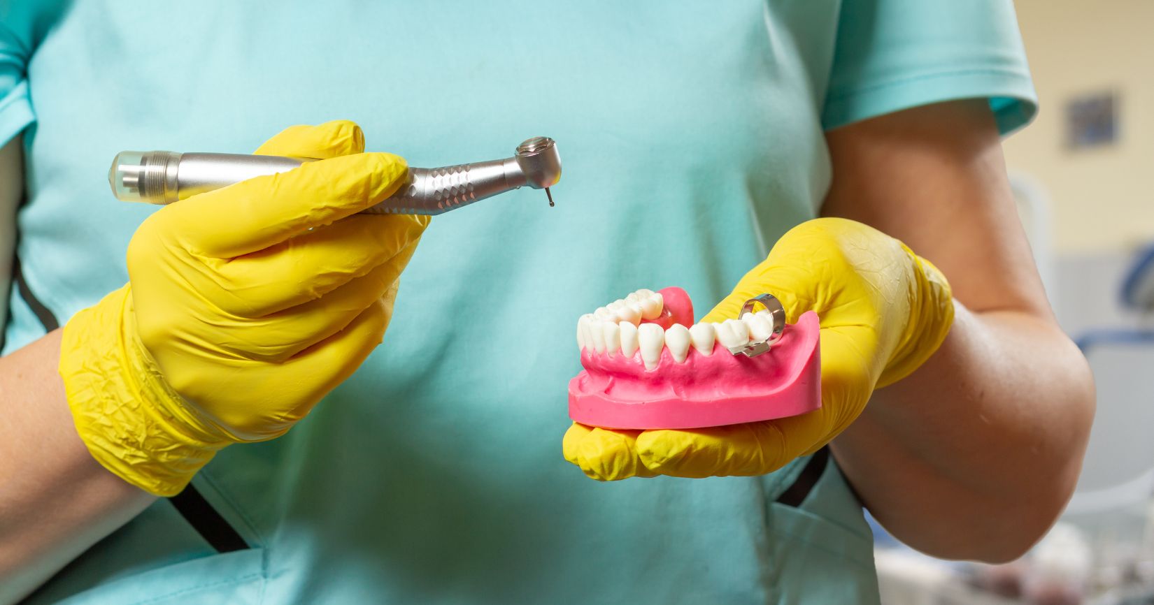 Dental professional in teal scrubs and yellow gloves holds a dental drill and a pink jaw model with implant framework in a clinic setting