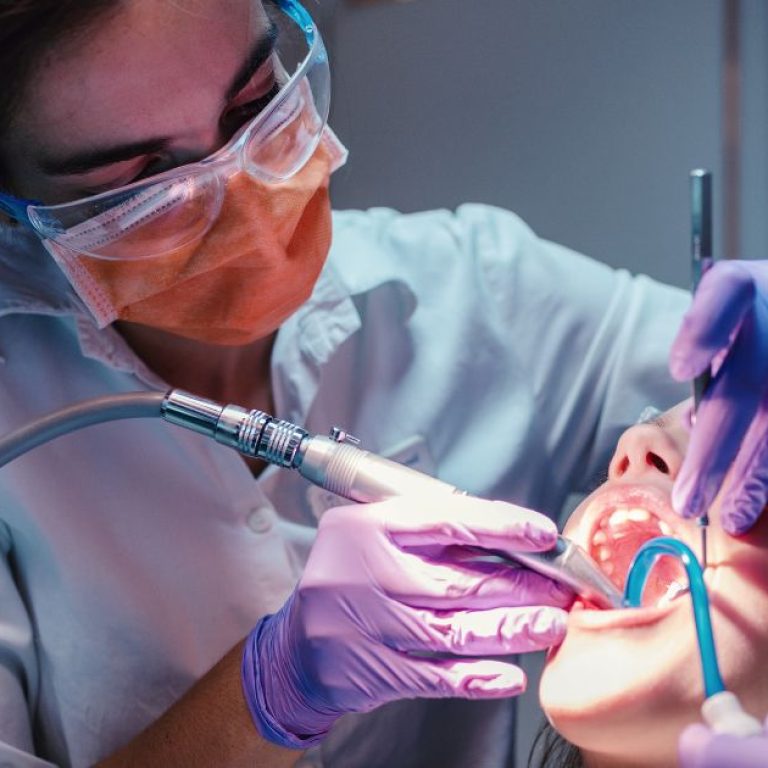 Dentist in protective gear performing a dental procedure using a drill on a patient&rsquo;s open mouth while wearing gloves and safety glasses.